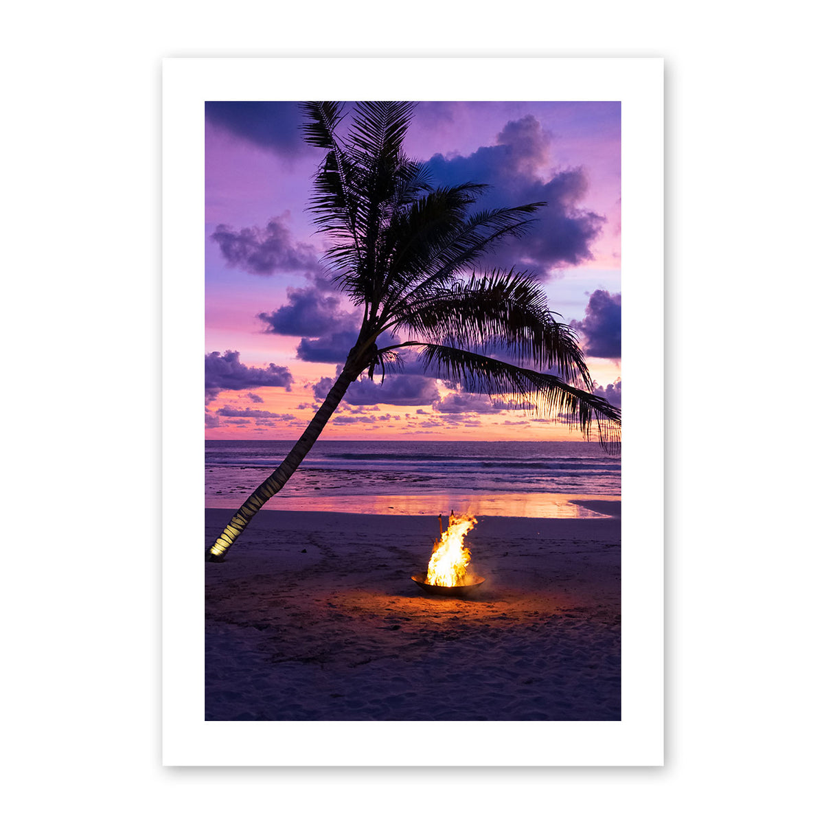 Photo of a fire pit by a palm tree on a beach with a sunset sky.